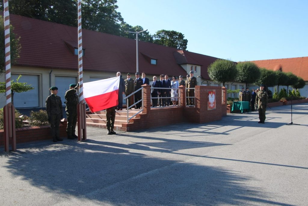 Grupa osób oraz żołnierzy na placu jednostki wojskowej. Na maszt wciągana jest flaga w barwach biało-czerwonych.