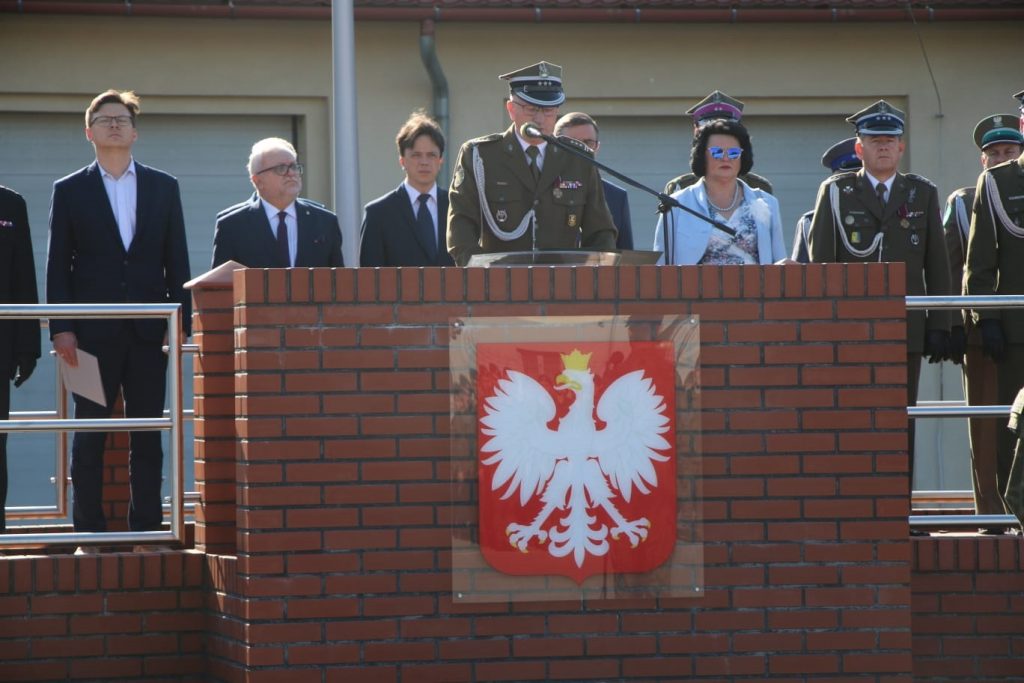 Mężczyzna w mundurze przemawia do mikrofonu. W tle grupa osób stojących obok siebie.