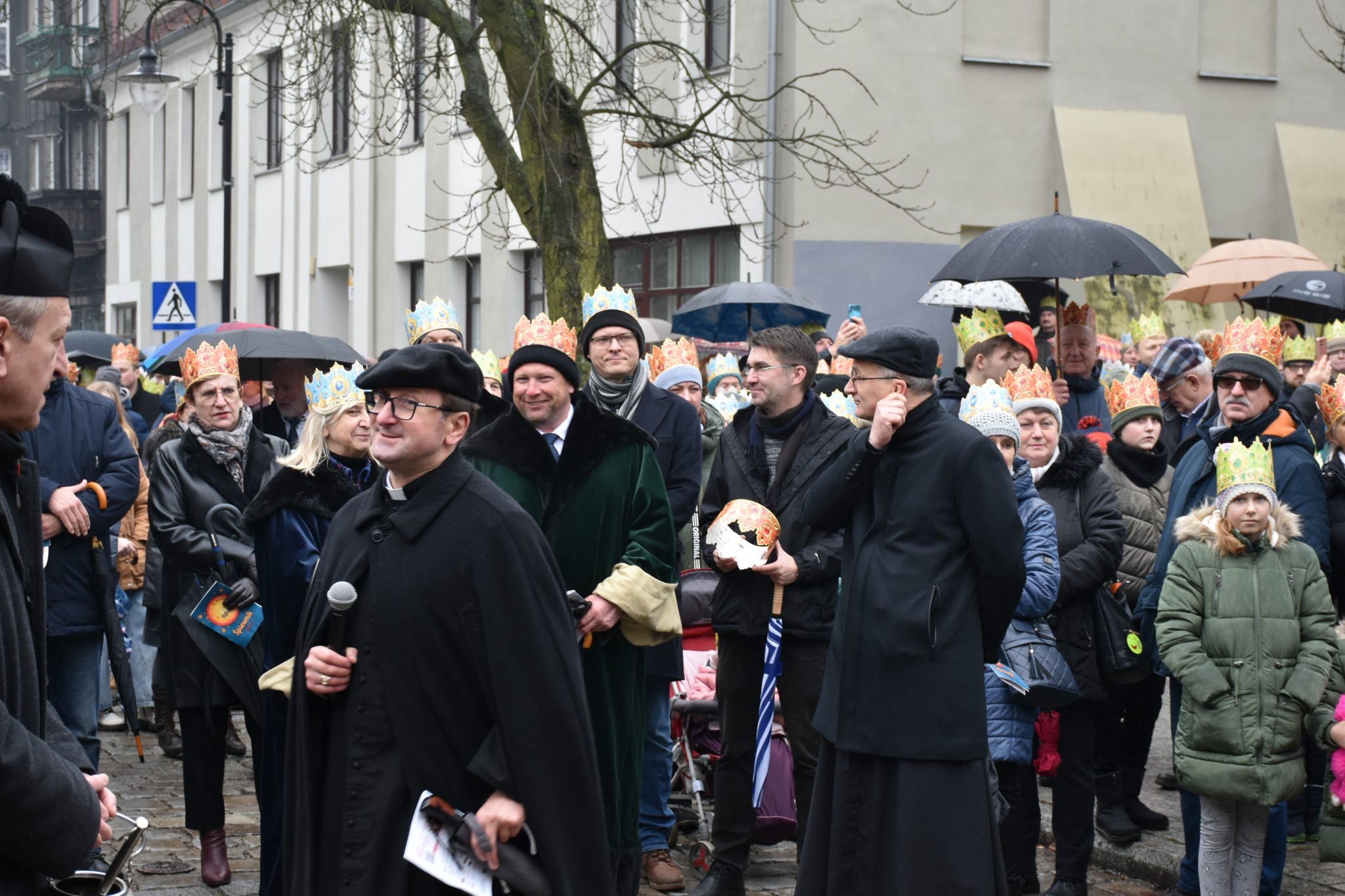 Duża grupa osób, w tle wysoki budynek. Część osób trzyma w rękach parasole.