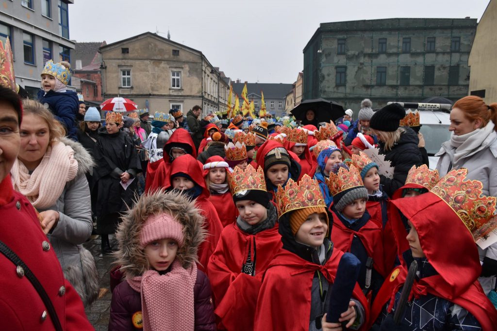 Grupa dzieci w czerwonych pelerynach i kartonowych czapeczkach w kształcie korony. W tle duża grupa osób i wysoka zabudowa.