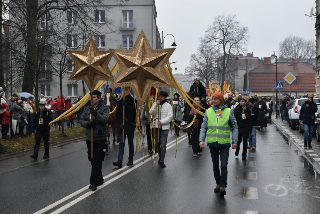 Duża grupa osób przemieszczająca się drogą. Na pierwszym planie osoby niosące dekoracje w kształcie gwiazd.