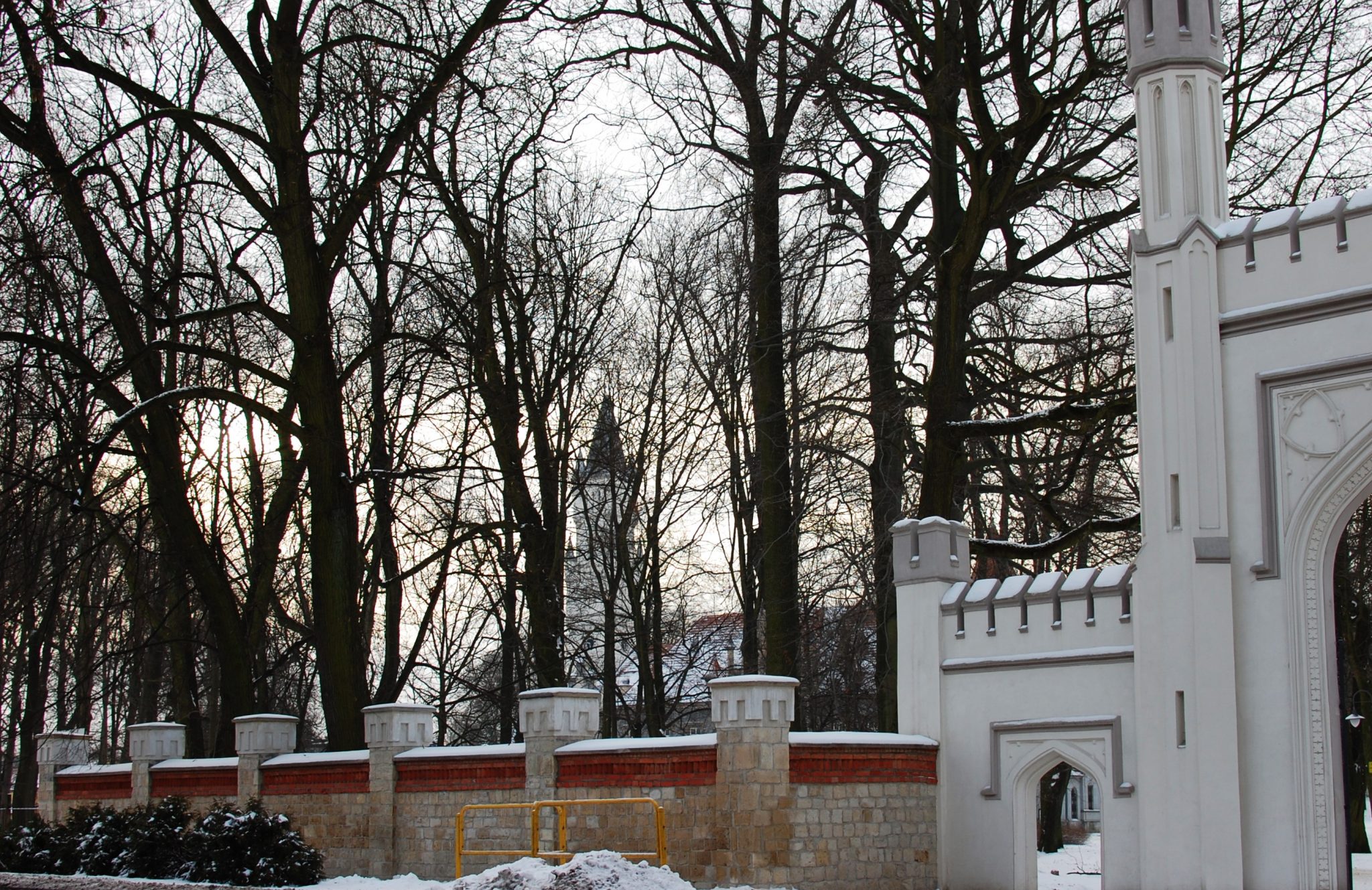 Ogrodzenie kamienne z ozdobną bramą wjazdową. W tle widoczne wysokie korony drzew bez liści oraz wysoka wieża.