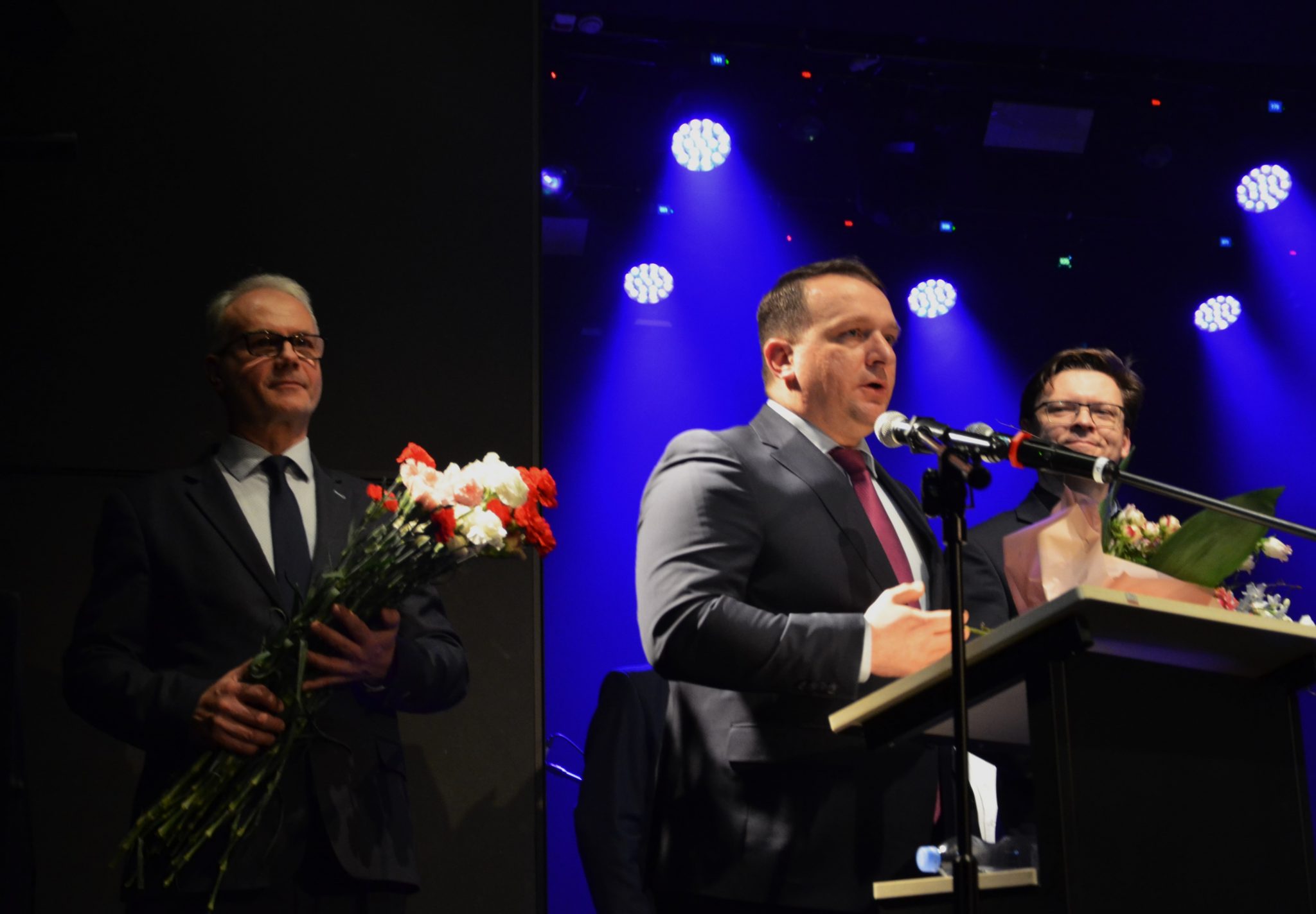 Mężczyzna podczas przemowy do mikrofonu. Po lewej stronie mężczyzna z bukietem biało-czerwonych kwiatów.