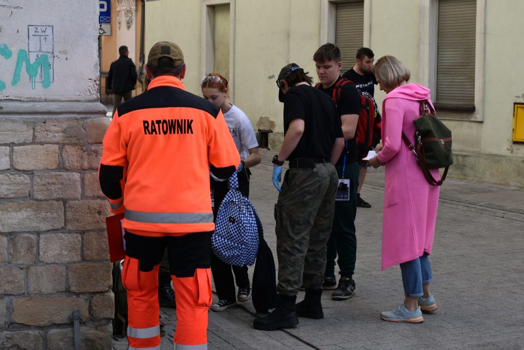 Grupa osób na ulicy. Na pierwszym planie osoba odwrócona plecami w pomarańczowym kombinezonie z napisem ratownik.