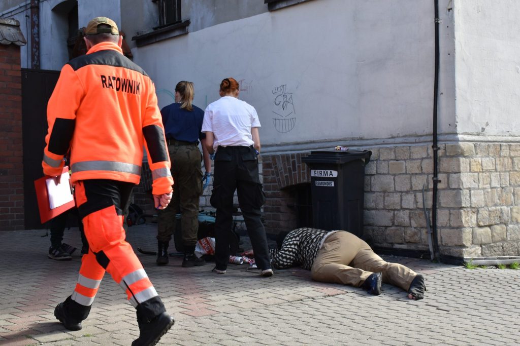 Grupa osób na ulicy. Na pierwszym planie osoba odwrócona plecami w pomarańczowym kombinezonie z napisem ratownik. W tle osoba leżąca na chodniku pod ścianą budynku.