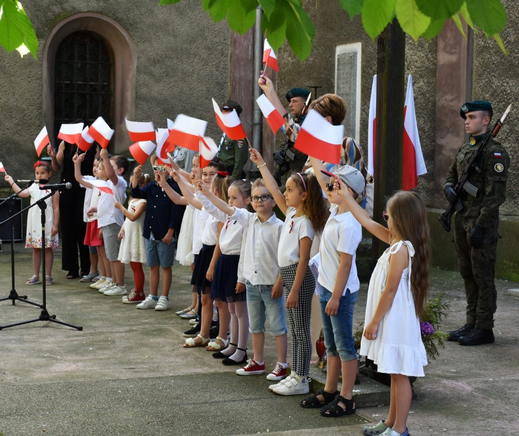 Grupa dzieci stojących przed budynkiem. Wszystkie dzieci trzymają w rękach biało-czerwone flagi.