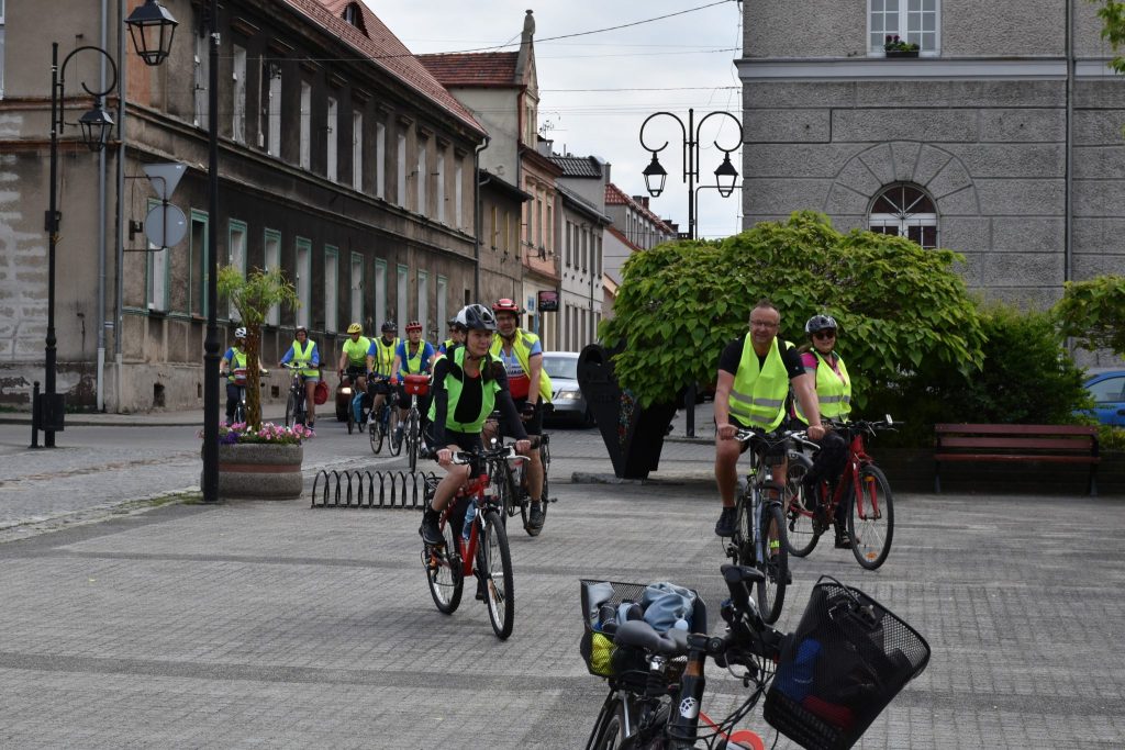 Grupa rowerzystów w kamizelkach jedzie uliczką pomiędzy budynkami.