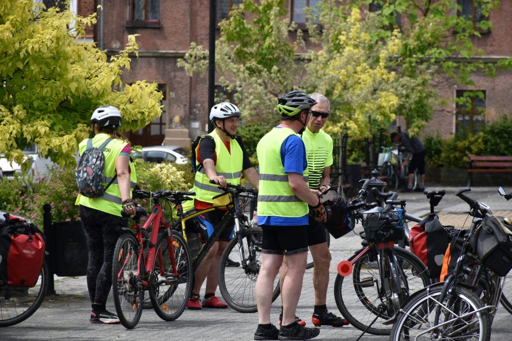Grupa osób znajdująca się przy rowerach, w tle zieleń i zabudowania.