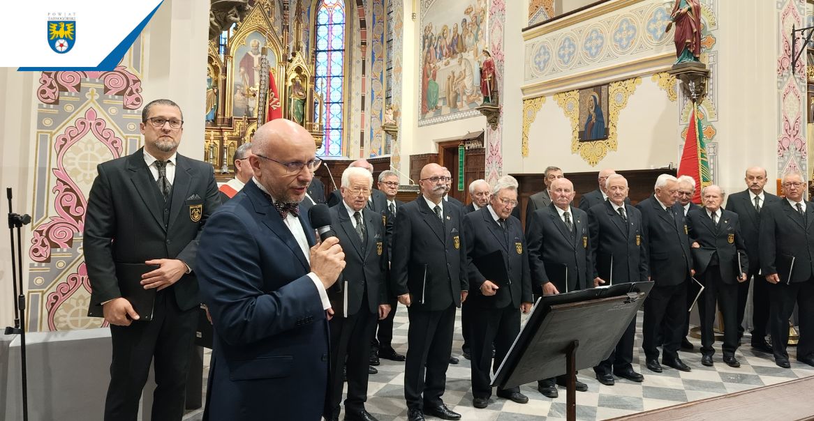 Grupa mężczyzn stoi w kościele. Wszyscy są ubrani w garnitur. Na pierwszym planie przemawia konferansjer.