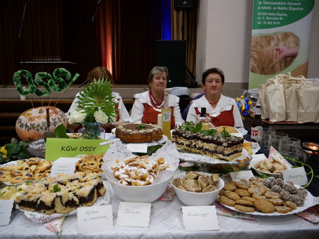 na pierwszym planie stół, na stole dużo ciast i ozdób, za stołem siedzą trzy kobiety, ubrane w regionalne stroje, w tle scena, po prawej stół z torbami, za stołem roll-up w tle scena, po prawej stół z torbami, za stołem roll-up