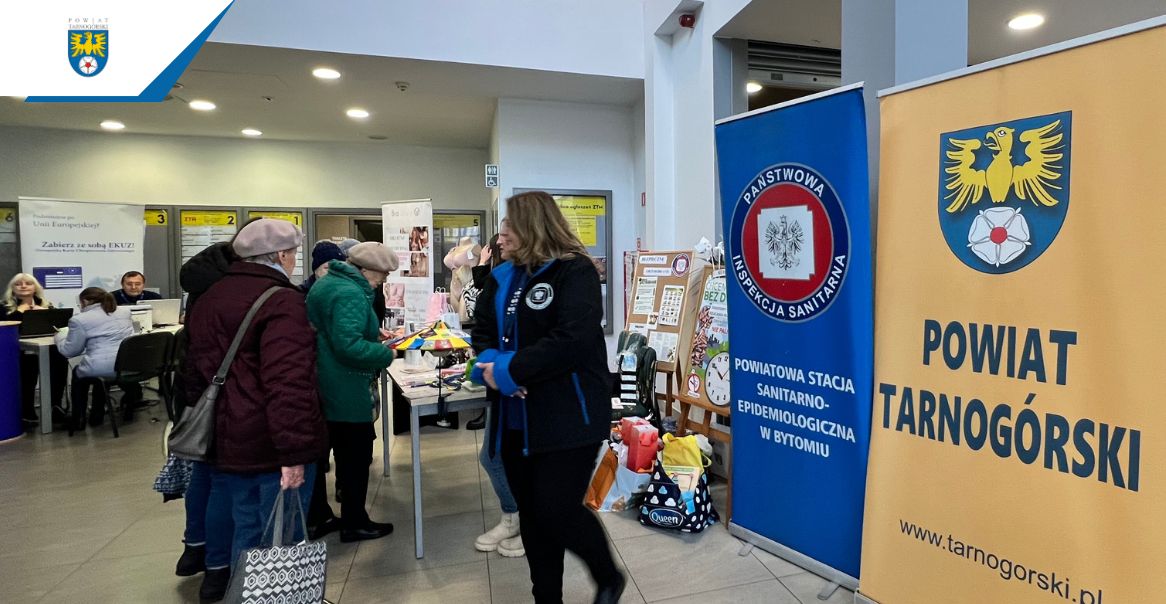 Zdjęcie przedstawiające ludzi w tle roll up w kolorze żółtym z herbem Powiatu Tarnogórskiego oraz drugi niebieski z logiem Sanepidu