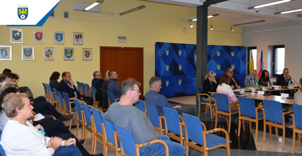 Sala sesyjna pełna siedzących ludzi. W tle ścianka z herbami Powiatu Tarnogórskiego.