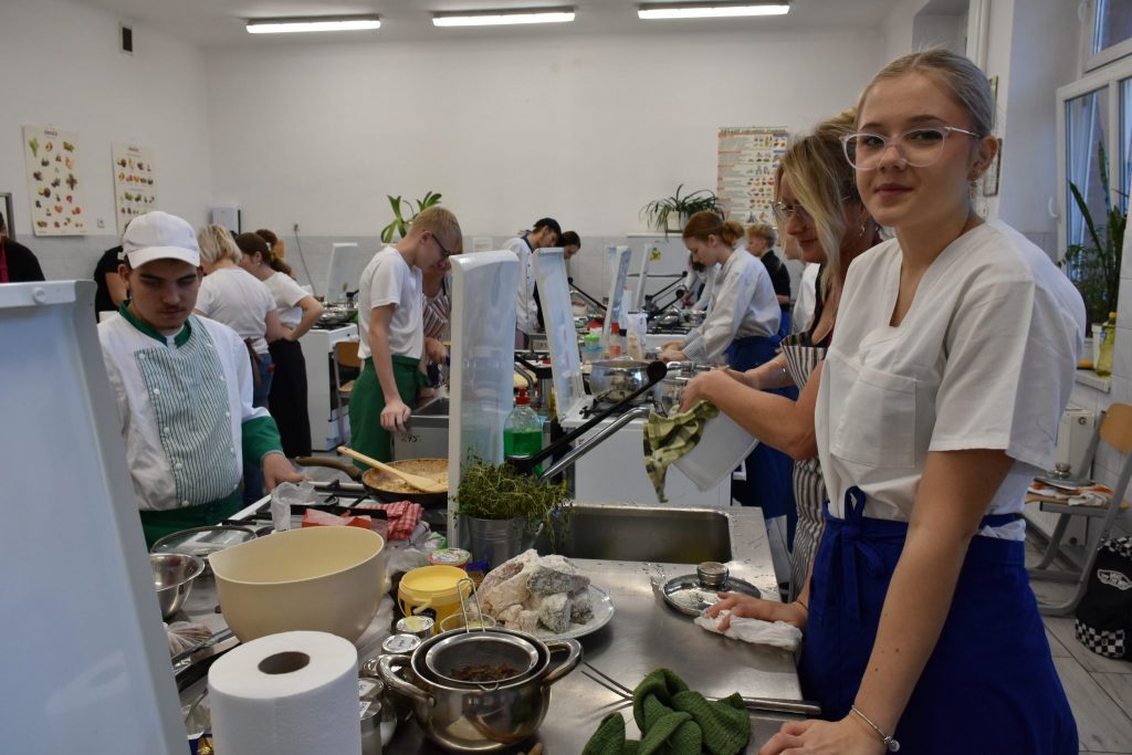 pomieszczenie kuchnia, wiele stanowisk do gotowania, różne produkty spożywcze na blatach kuchennych, kobiety i mężczyźni gotują