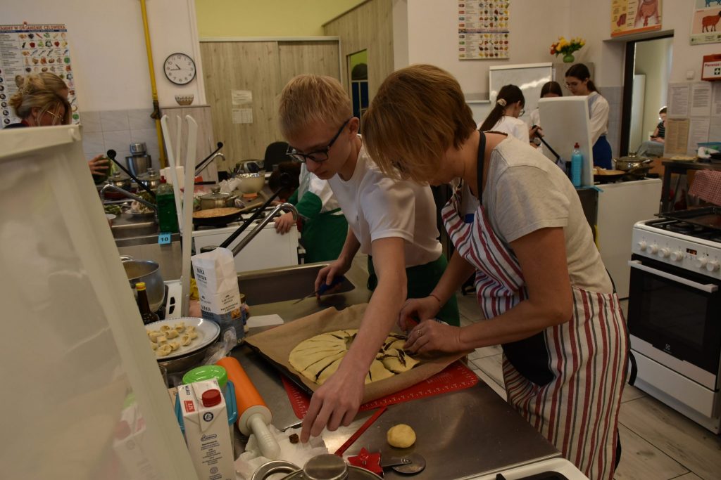 pomieszczenie kuchenne, młody mężczyzna oraz kobieta stoją przy blacie kuchennym, gotują, na blacie wiele przyrządów kuchennych, w tle inne osoby gotujące