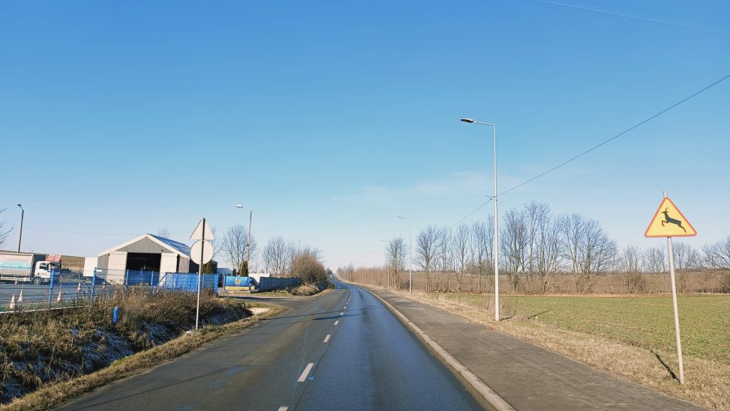 pośrodku droga z linią przerywaną, po prawej chodnik i fragment pola oraz latarnia uliczna, po lewej rów, znak pionowy oraz w tle budynek z otwartą bramą