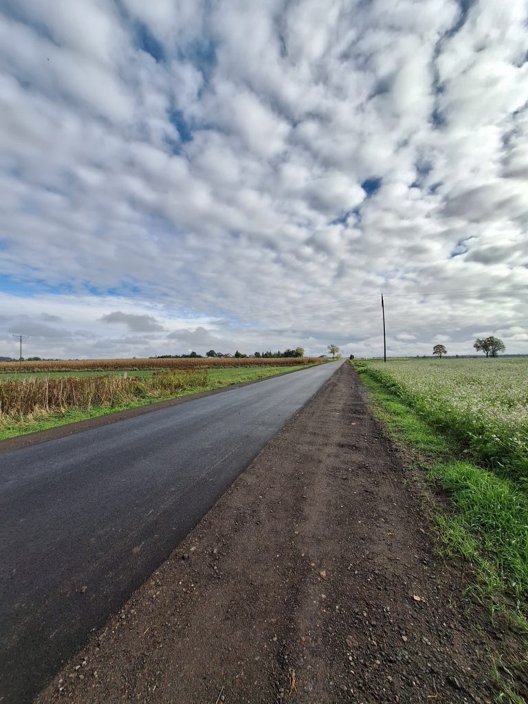 na górze pierzaste chmury, niebo niebieskie, poniżej w głębi daleki horyzont drogi, pusta droga, po prawej pole, a w tle biały dom, po lewe pole, w oddali drzewa