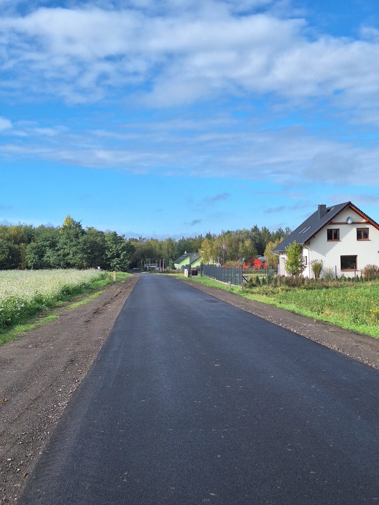 na górze błękitne niebo, poniżej pasmo drzew i droga, po prawej fragment pola oraz biały dom, po lewej fragment pola, w oddali drzewa