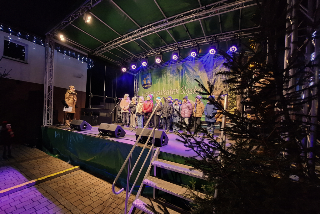 na pierwszym planie po prawej choinka, obok schody i wejście na scenę, na scenie stoją w dwóch rzędach dzieci, po lewej kobieta, w tle bilboard i światła