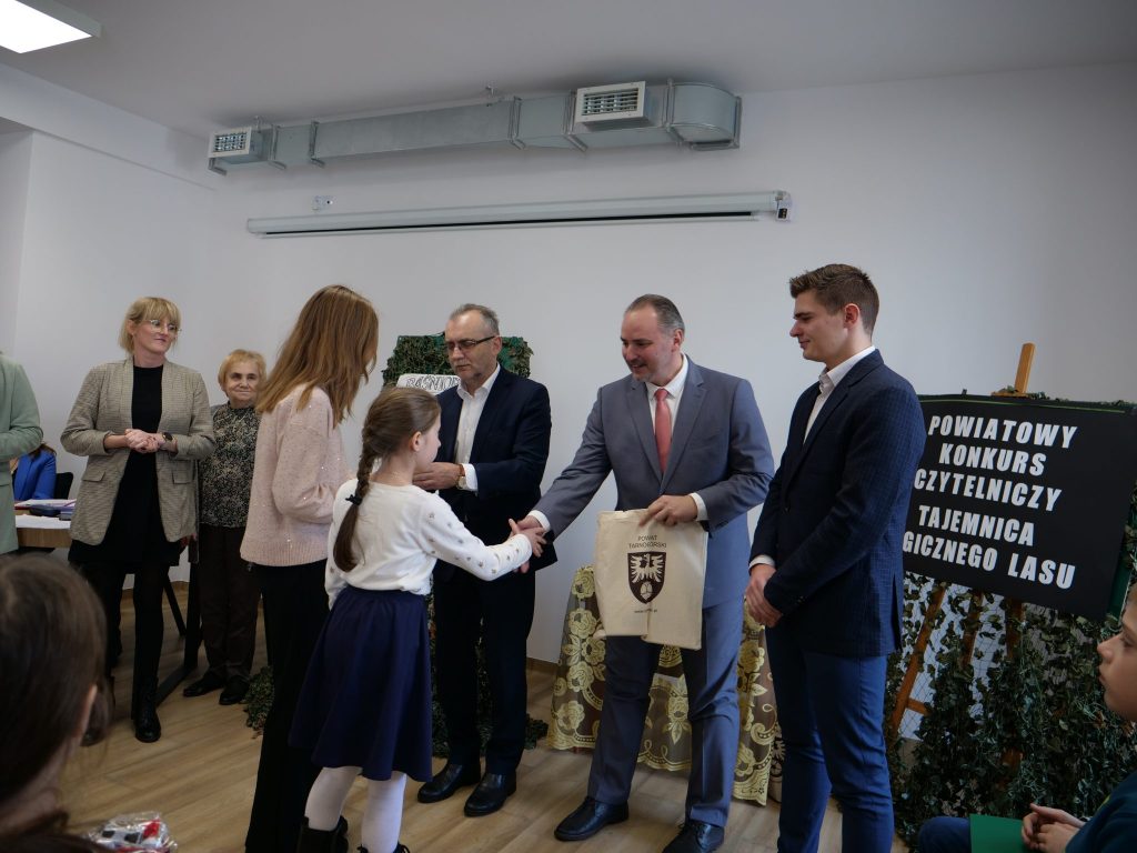 dorośli stoją przodem, dzieci stoją tyłem, dostają torbę, po prawej napis na czarnym tle, na sztaludze 