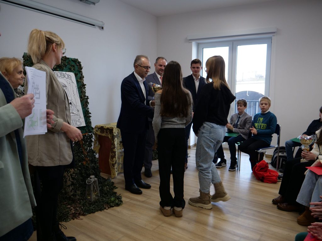 dorośli stoją przodem, dzieci stoją tyłem, niektóre siedzą, w tle okno, dookoła białe ściany 