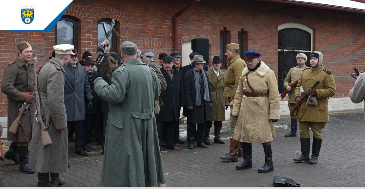 Grupa mężczyzn stojących przed budynkiem. Ubrani są w stroje dawnych czasów. W rekach trzymają broń.