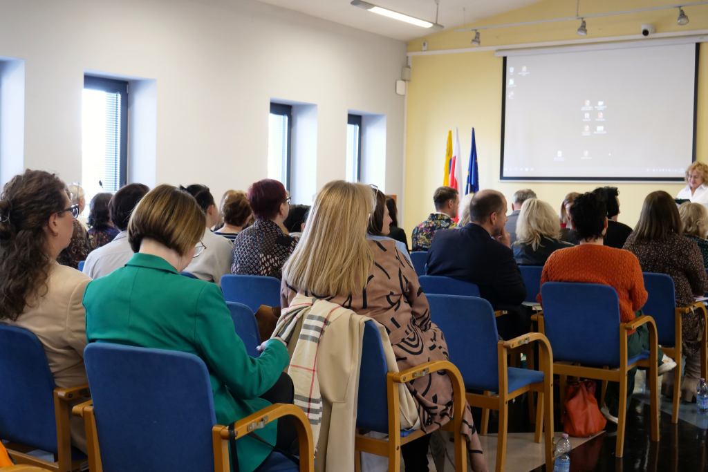 publiczność, kobiety i mężczyźni siedzą na krzesłach, zwróceni tyłem, w tle na ścianie wyświetlacz