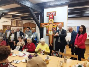 Spotkanie w sali z dziełami sztuki i dużym krzyżem z ikoną Chrystusa w tle. Seniorzy siedzą przy stole, haftują i piją kawę, a za nimi stoją goście, w tym osoby w strojach kolarskich oraz elegancko ubrany mężczyzna i kobieta. Panuje serdeczna i ciepła atmosfera.