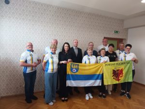 Grupowe zdjęcie osób ubranych w białe koszulki kolarskie z niebiesko-żółtym pasem i herbem, stojących na tle jasnej ściany w pomieszczeniu. Dwie osoby w środku (kobieta i mężczyzna) ubrane są elegancko. Część grupy trzyma dwie flagi – jedna z herbem i napisami w języku polskim, druga zielono-żółta z napisem „Powiat Tucholski”.