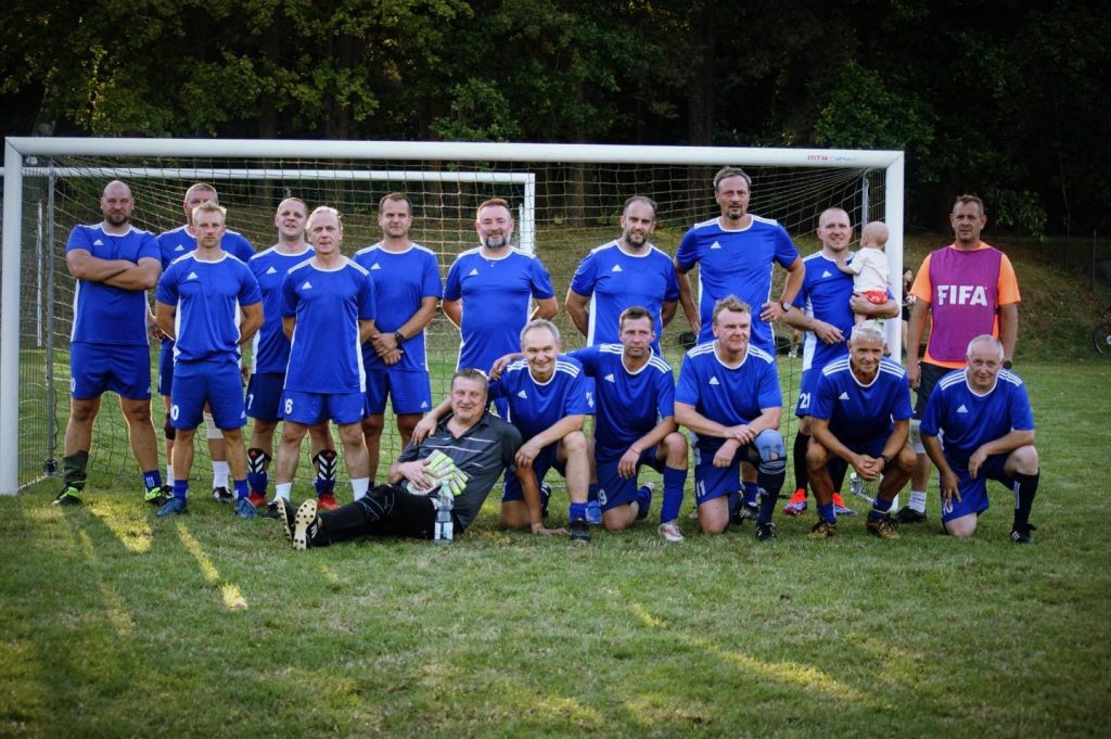 cała drużyna piłkarzy, w tych samych strojach sportowych, pozuje do zdjęcia na boisku, w tle bramka piłkarska 