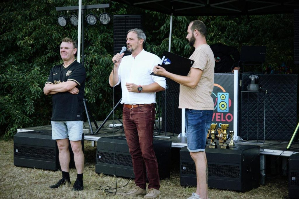 trzech mężczyzn, jeden ubrany w koszulkę piłkarską, obok stoi mężczyzna który trzyma w ręku mikrofon, po prawej mężczyzna który trzyma w ręku teczkę, jest ubrany w koszulkę i krótkie spodenki