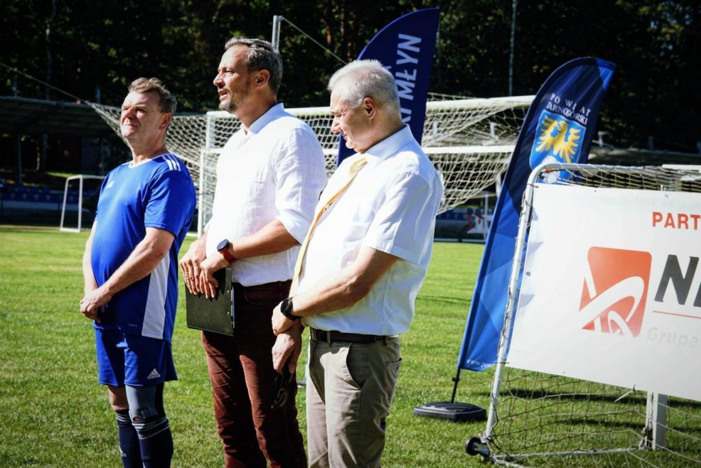 trzech mężczyzn stoi na boisku, jeden ubrany na sportowo, dwóch w stroje formalne