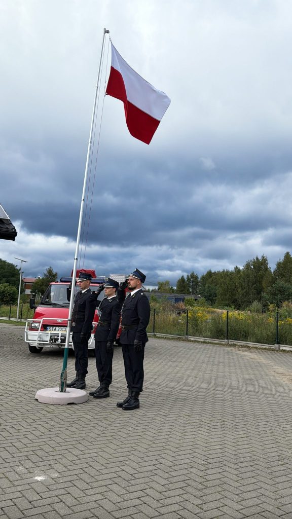 stoi trzech mężczyzn w mundurach, obok maszt z flagą polski, za nimi wóz strażacki, w tle ogrodzenie i zielone krzewy i drzewa, powyżej zachmurzone niebo