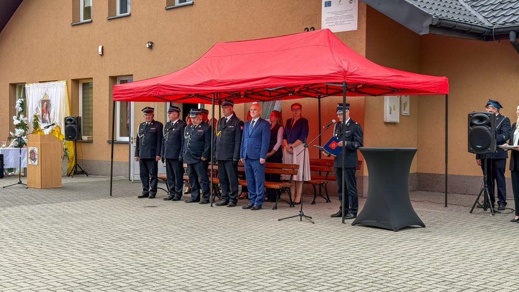 z przodu kostka brukowa, po prawej głośnik, za głośnikiem mężczyzna w mundurze i fragment kobiety, na środku ludzie stoją pod namiotem, mężczyzna w mundurze który czyta, mówi do mikrofonu, po lewej mównica , w oddali zdjęcie papieża, w tle budynek z oknami.