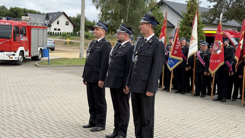 na środku stoi trzech mężczyzn, po prawej mężczyźni trzymają sztandary, po lewej stoi wóz strażacki, za nim fragment samochodu osobowego, w tle zabudowania i drzewa, powyżej fragment zachmurzonego nieba