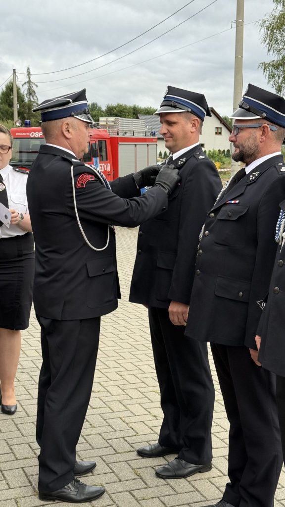 na pierwszym planie stoi trzech mężczyzn w mundurze, po prawej fragment mężczyzny w mundurze, po prawej kobieta, w tle wóz strażacki, drzewa i zabudowania, powyżej słupy energetyczne z kablami i zachmurzone niebo