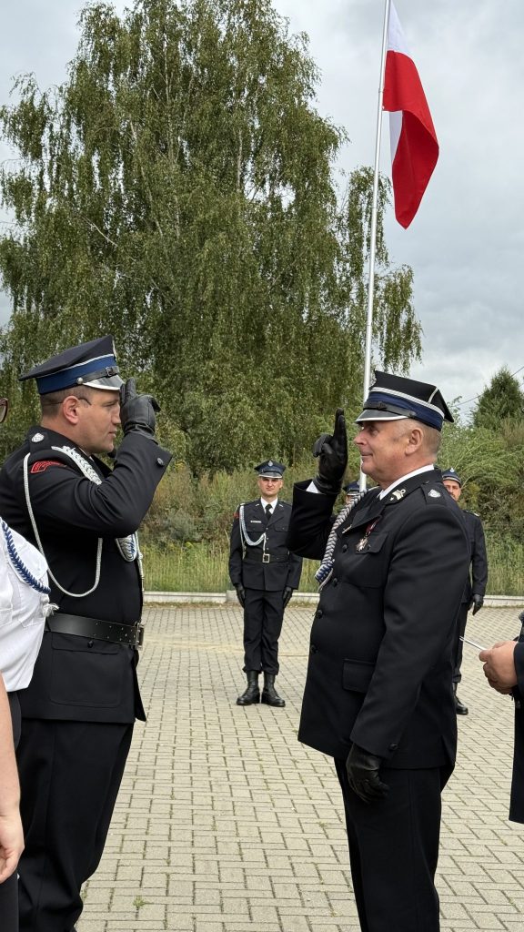 na pierwszym planie dwóch mężczyzn w mundurach, stoją na kostce brukowej, na drugim planie mężczyzna, powyżej fragment masztu z flagą polski, w tle drzewa