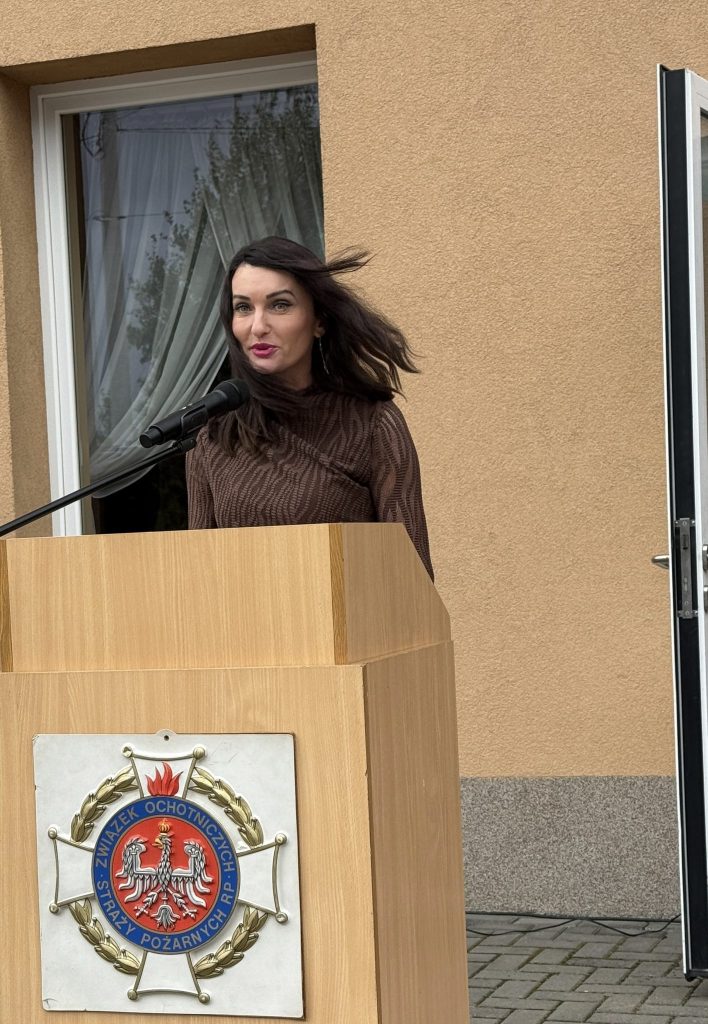 na pierwszym planie mównica, za nią kobieta mówiąca do mikrofonu, w tle fragment budynku i okno