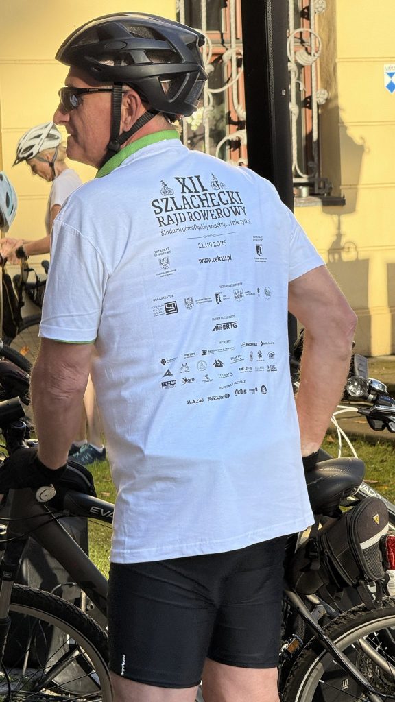 mężczyzna, ubrany na sportowo , na głowie ma kask, stoi przy rowerze, w tle inni uczestnicy