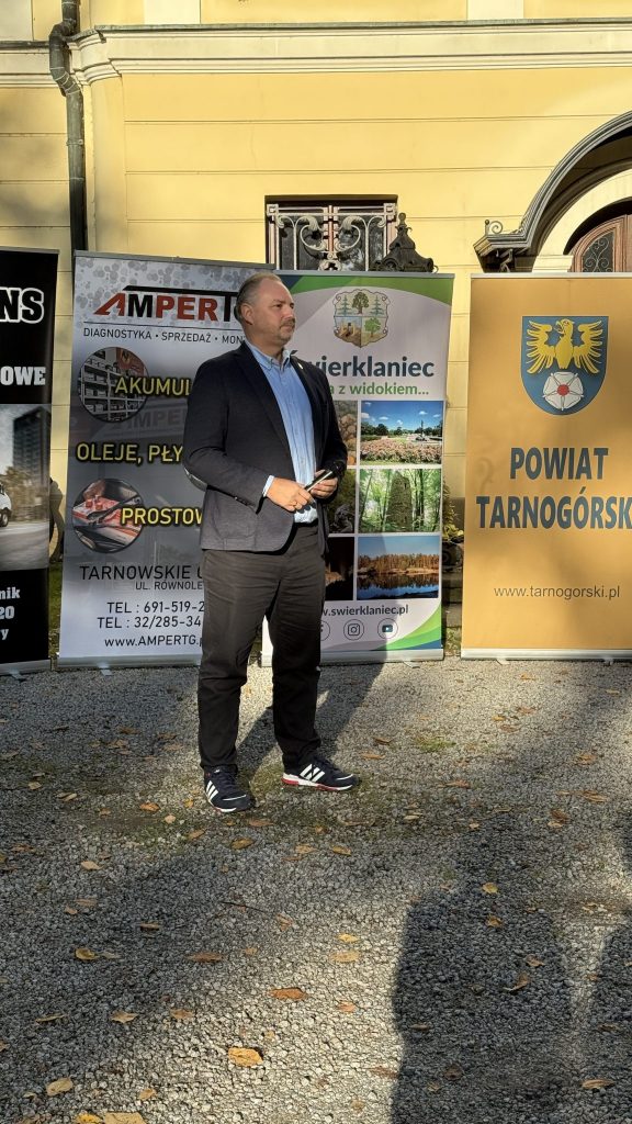 mężczyzna, ubrany półformalnie, stoi na drodze utwardzonej kamieniami, w tle kolorowe banery, za nimi fragment pałacu