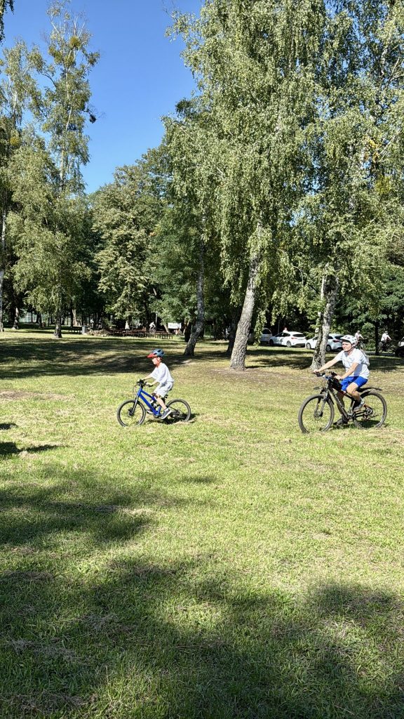 dwójka dzieci, na rowerach, na głowie kaski, jadą po trawie rowerami, w tle wysokie drzewa