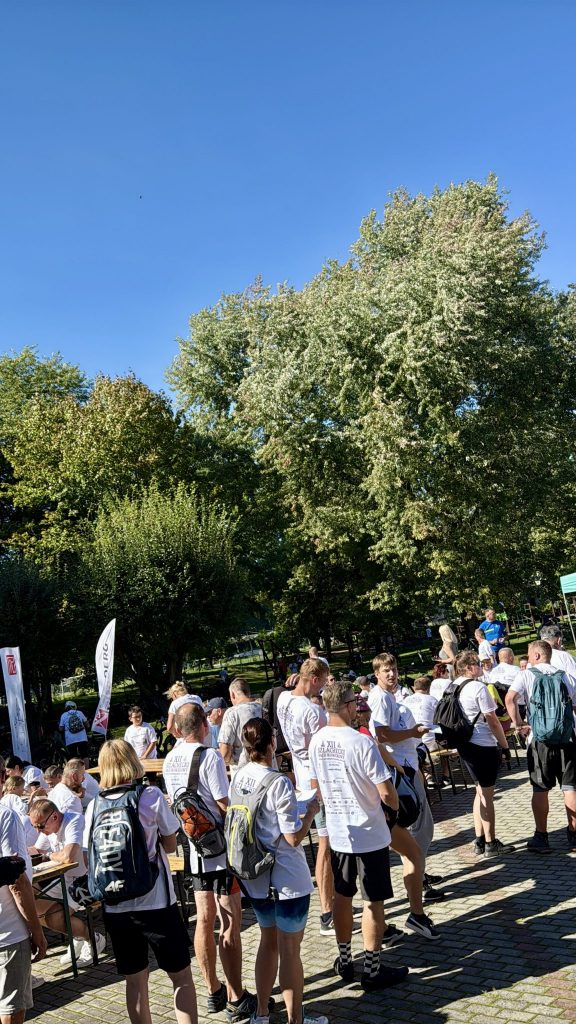 grupa osób, jedni stoją , drudzy siedzą na ławkach przy stołach, wszyscy ubrani na sportowo, niektórzy maja na plecach plecaki