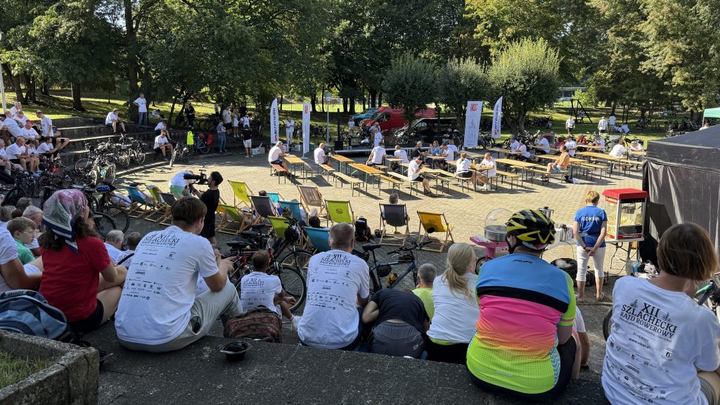 uczestnicy w sportowych strojach, siedzą na ławkach w amfiteatrze plenerowym, wokół stoją rowery, na środku ławki i stoły oraz leżaki , w tle drzewa