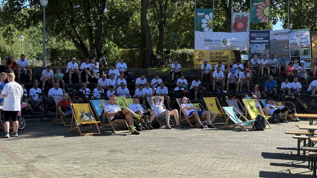 na pierwszym planie osoby siedzą na leżakach, za nimi duża ilość ludzi siedzi na ławkach amfiteatru plenerowego, w tle drzewa i kolorowe banery