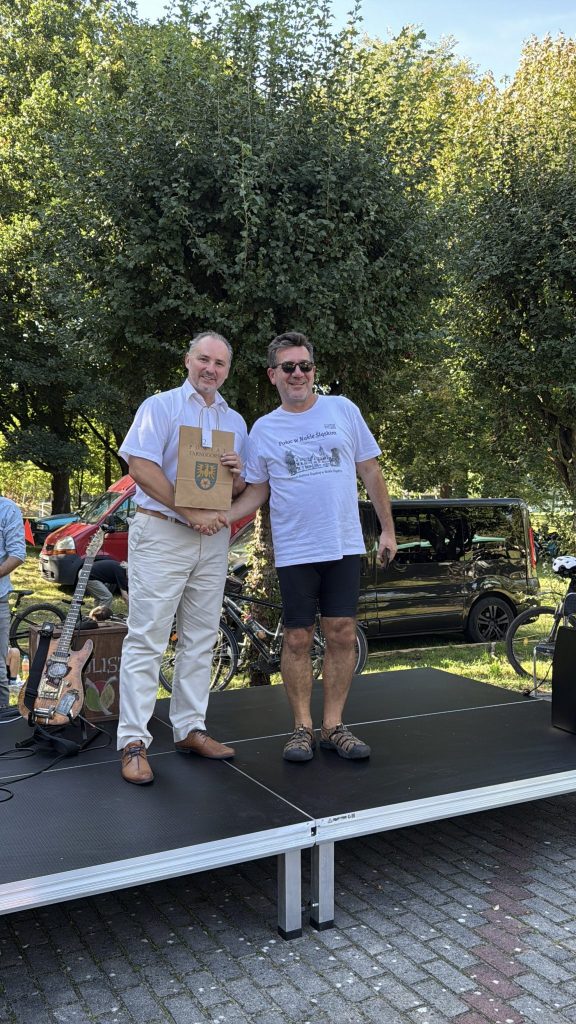dwóch mężczyzn, jeden ubrany elegancko trzyma w ręku upominek, drugi na sportowo, podają sobie ręce, stoją na scenie, w tle samochody, rowery i drzewa