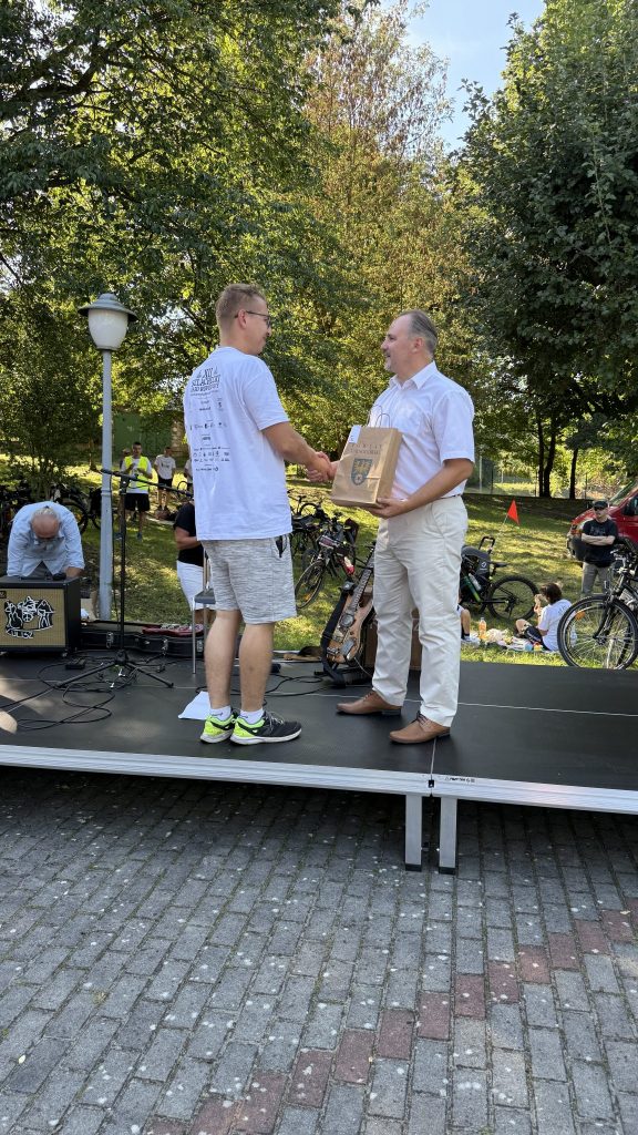 mężczyzna, elegancko ubrany, wręcza upominek mężczyźnie który jest ubrany na sportowo, stoją na scenie, wokół rowery, uczestnicy oraz drzewa