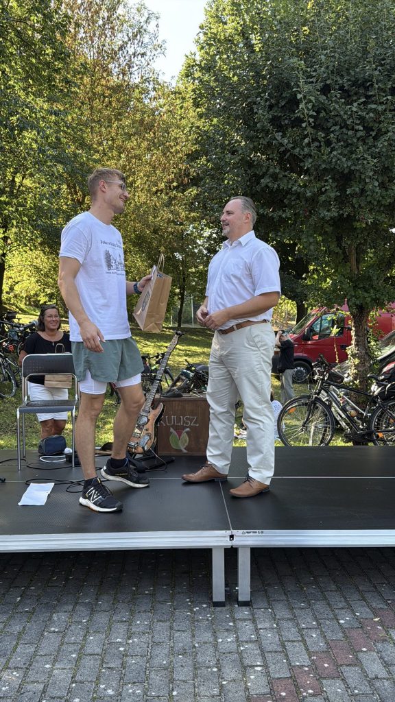 dwóch mężczyzn, uśmiechają się, jeden z nich trzyma w ręku upominek, za nim kobieta, uśmiecha się, w tle drzewa, rowery i samochody