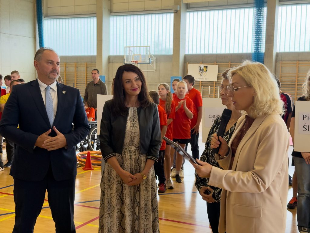 na pierwszym planie cztery dorosłe osoby, trójka kobiet i jeden mężczyzna, stoją w pomieszczeniu na hali sportowej, w tle dzieci ubrane na sportowo
