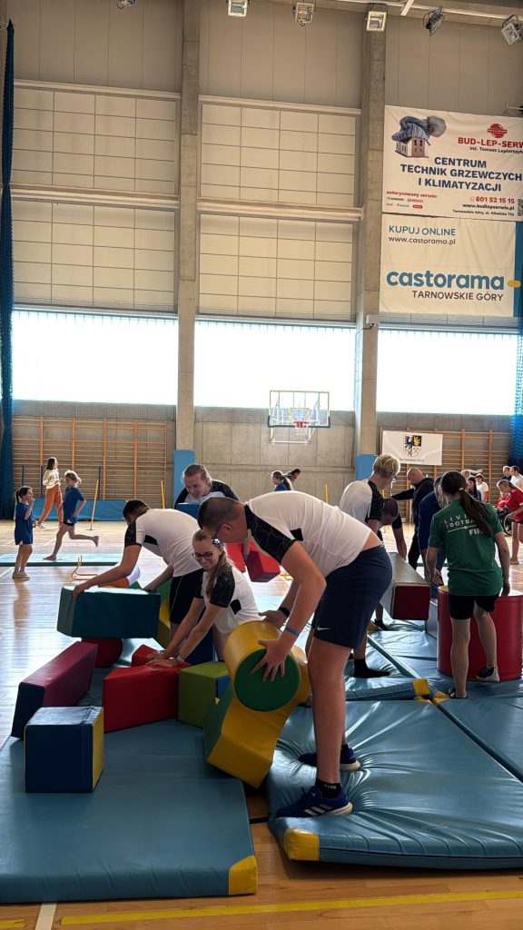hala sportowa, zawodnicy układają kolorowe piankowe klocki o różnych kształtach na materacach gimnastycznych, w tle inne osoby biorące udział w aktywnościach sportowych, w tle banery reklamowe i kosz do koszykówki