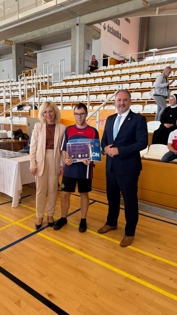 kobieta i mężczyzna oraz zawodnik sportowy z dyplomem w ręku, na hali sportowej, w tle trybuny 
