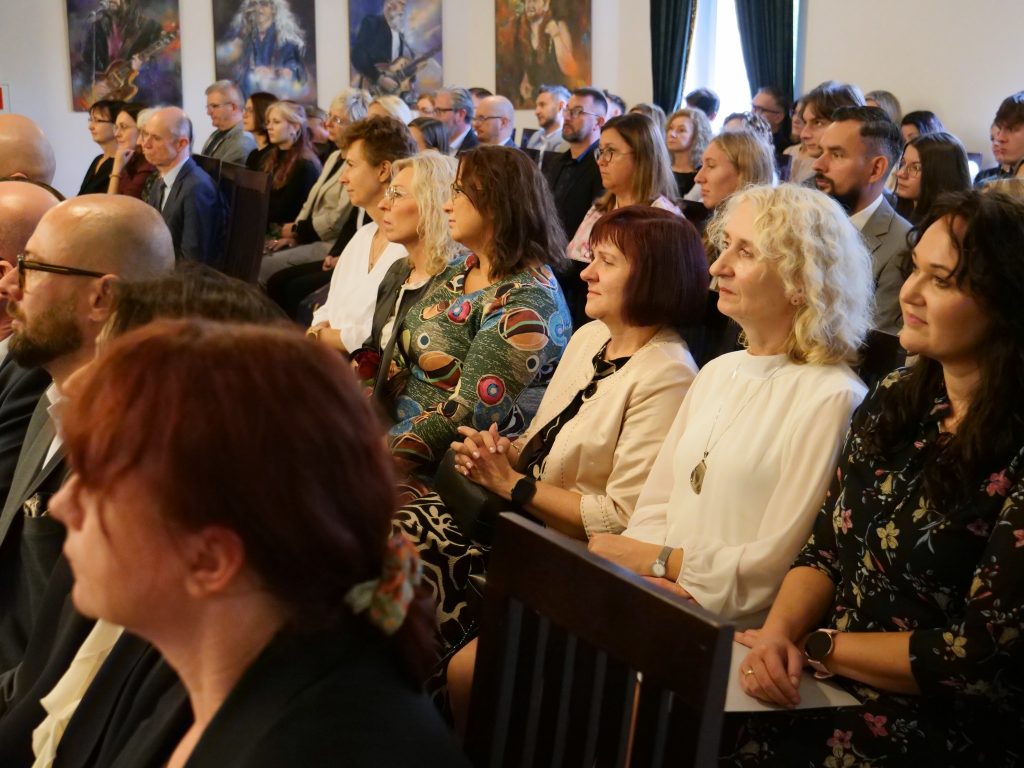 duża grupa osób siedzi na widowni, w tle na ścianie obrazy